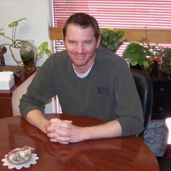 a man dressed in long sleeves, is sitting at a round wooden table that is so polished it  is reflecting his image. He is looking at the camera and smiling. Behind him are a variety of plants and a window with blinds on it.