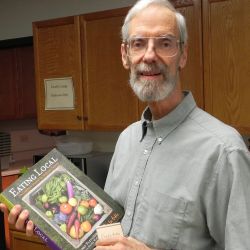 A picture of a man, standing in what appears to be a kitchen, he is wearing wire framed glasses and a button up polo. Looking directly into the camera he is holding a book labeled Eating Local. 